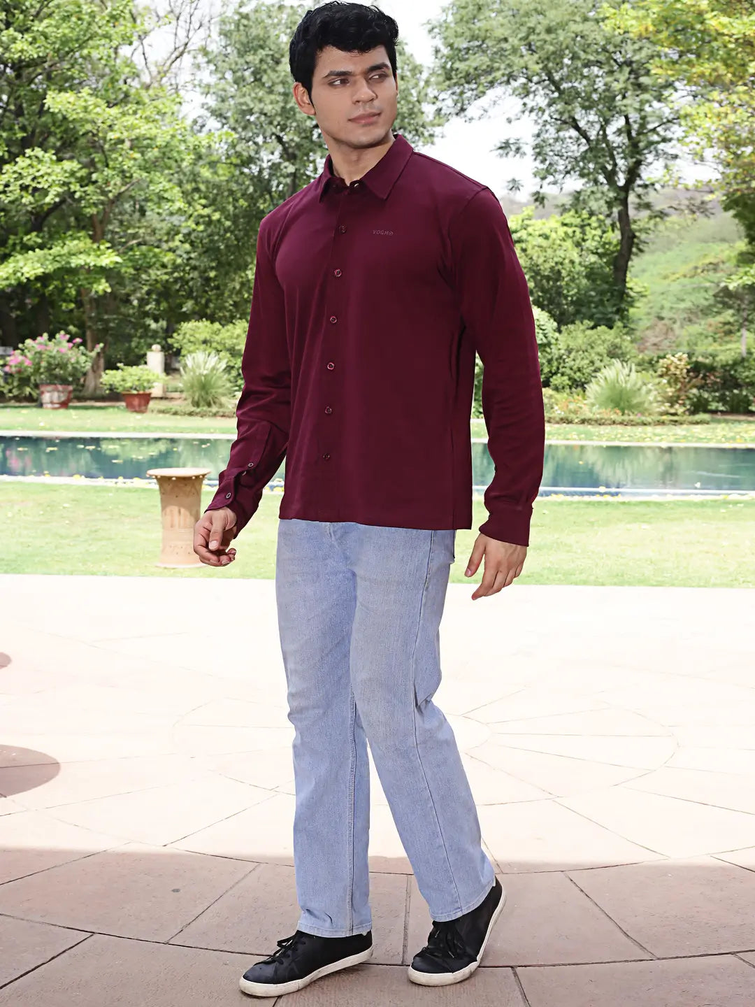Man wearing a maroon shirt and light blue jeans standing outdoors with greenery in the background