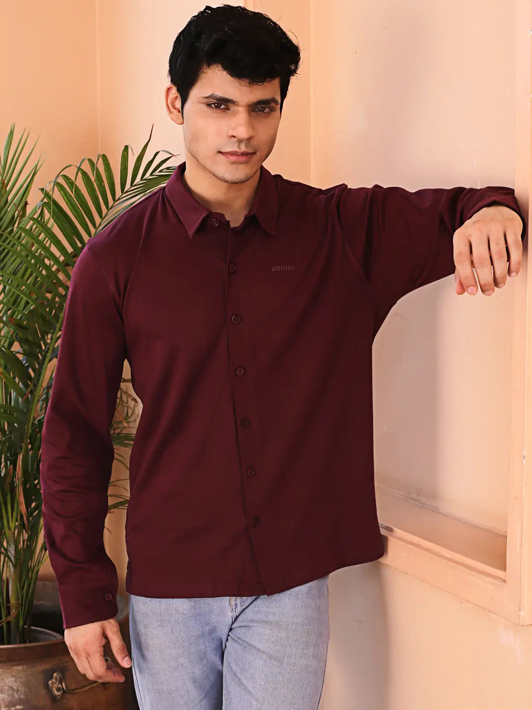 Man wearing a maroon shirt and light blue jeans standing indoors with a plant in the background