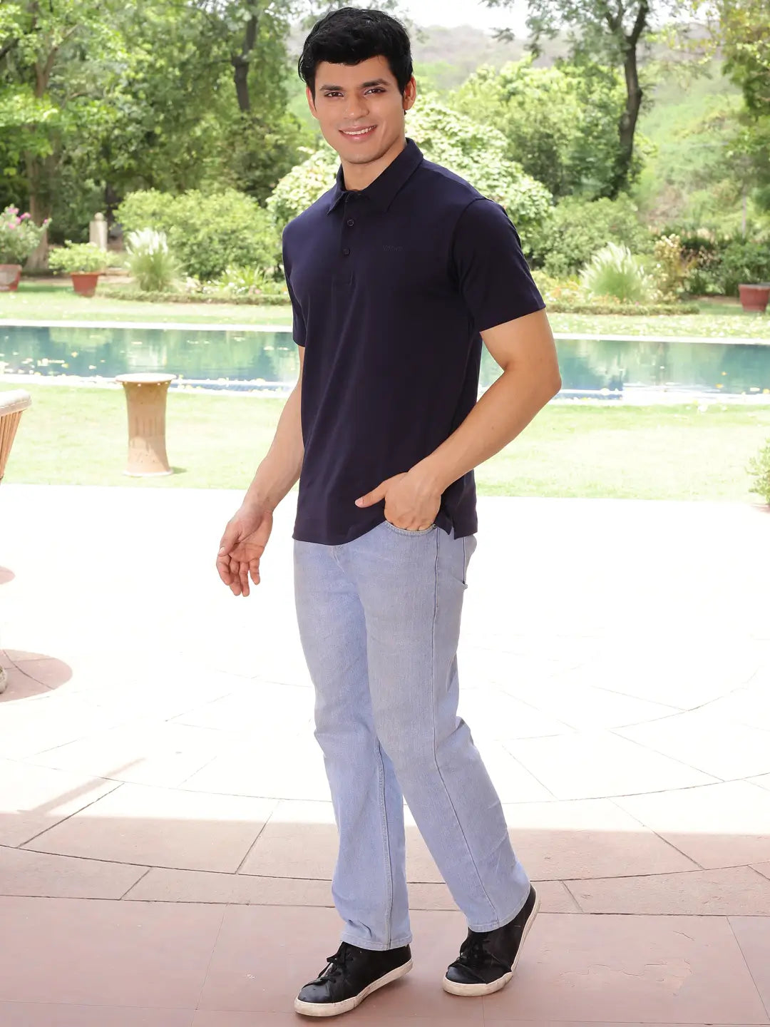 Man wearing a navy polo shirt and light blue jeans standing by a pool with greenery in the background