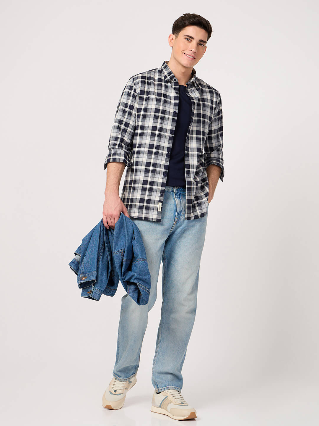 Man wearing a plaid shirt, blue jeans, and holding a denim jacket on a white background