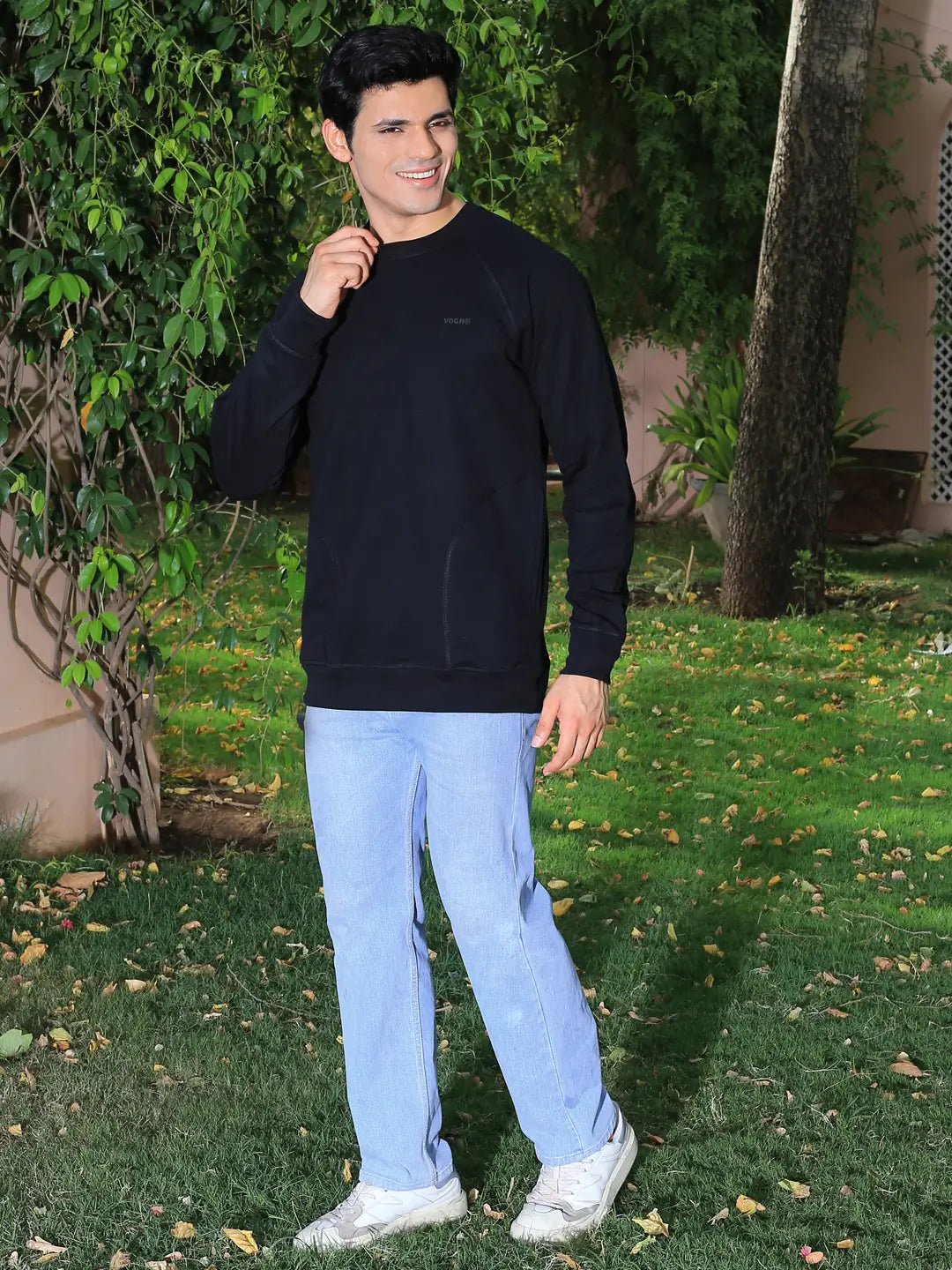 Man wearing a black sweatshirt and light blue jeans standing outdoors with greenery in the background