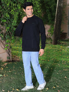 Man wearing a black sweatshirt and light blue jeans standing outdoors with greenery in the background
