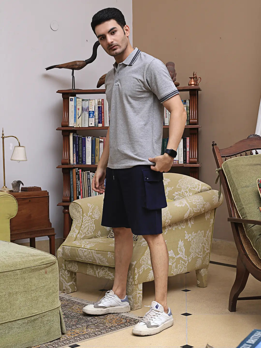 Man in gray polo shirt and navy shorts standing in a living room with bookshelf and furniture.