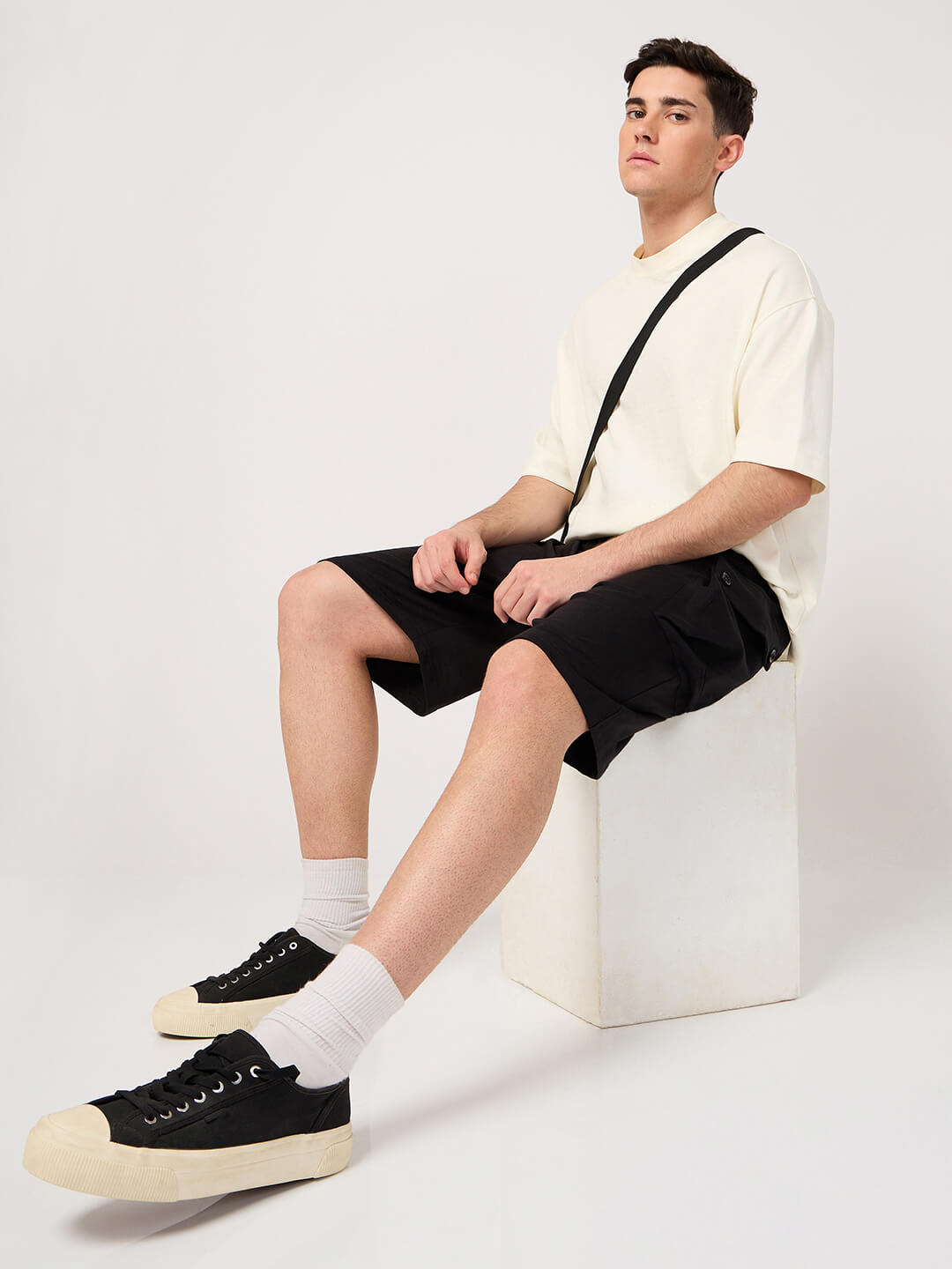 Person sitting on a white chair wearing a white shirt, black cargo shorts, and black sneakers with white socks.