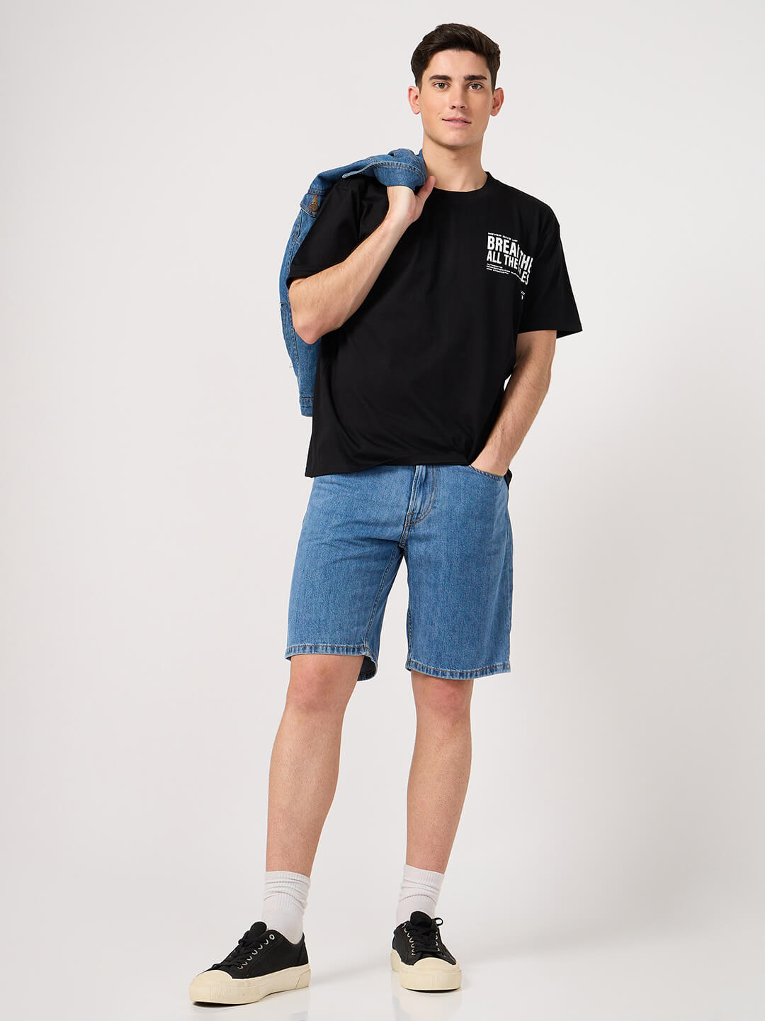 Man wearing a black printed t-shirt, blue denim shorts, and black sneakers on a white background