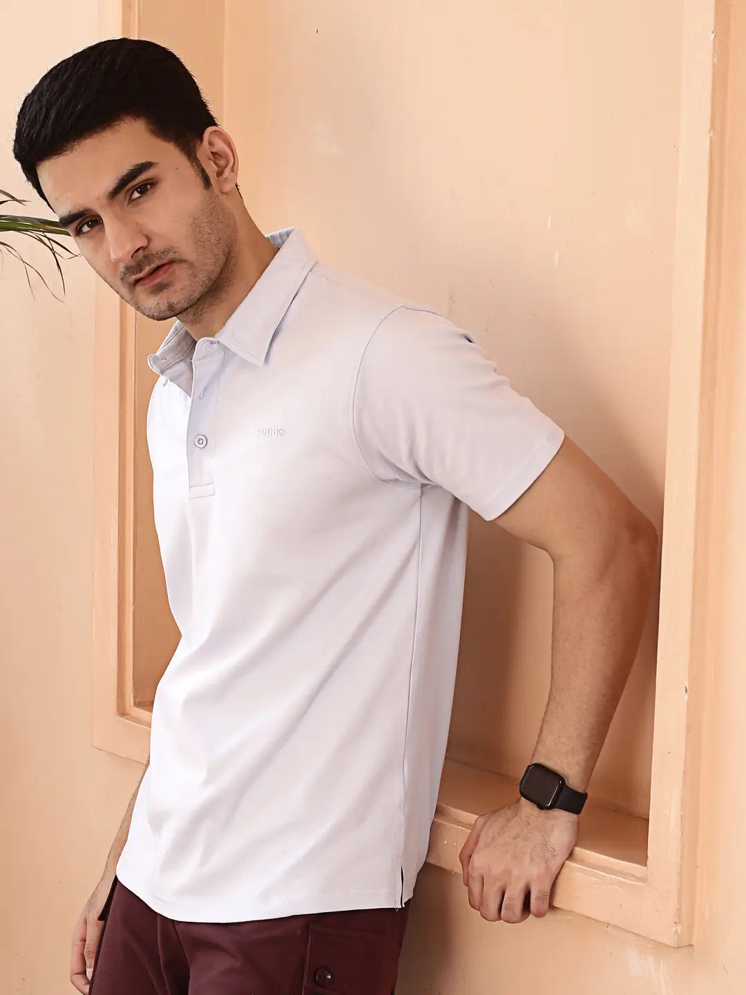 Man wearing a white polo shirt and maroon pants against a beige wall.