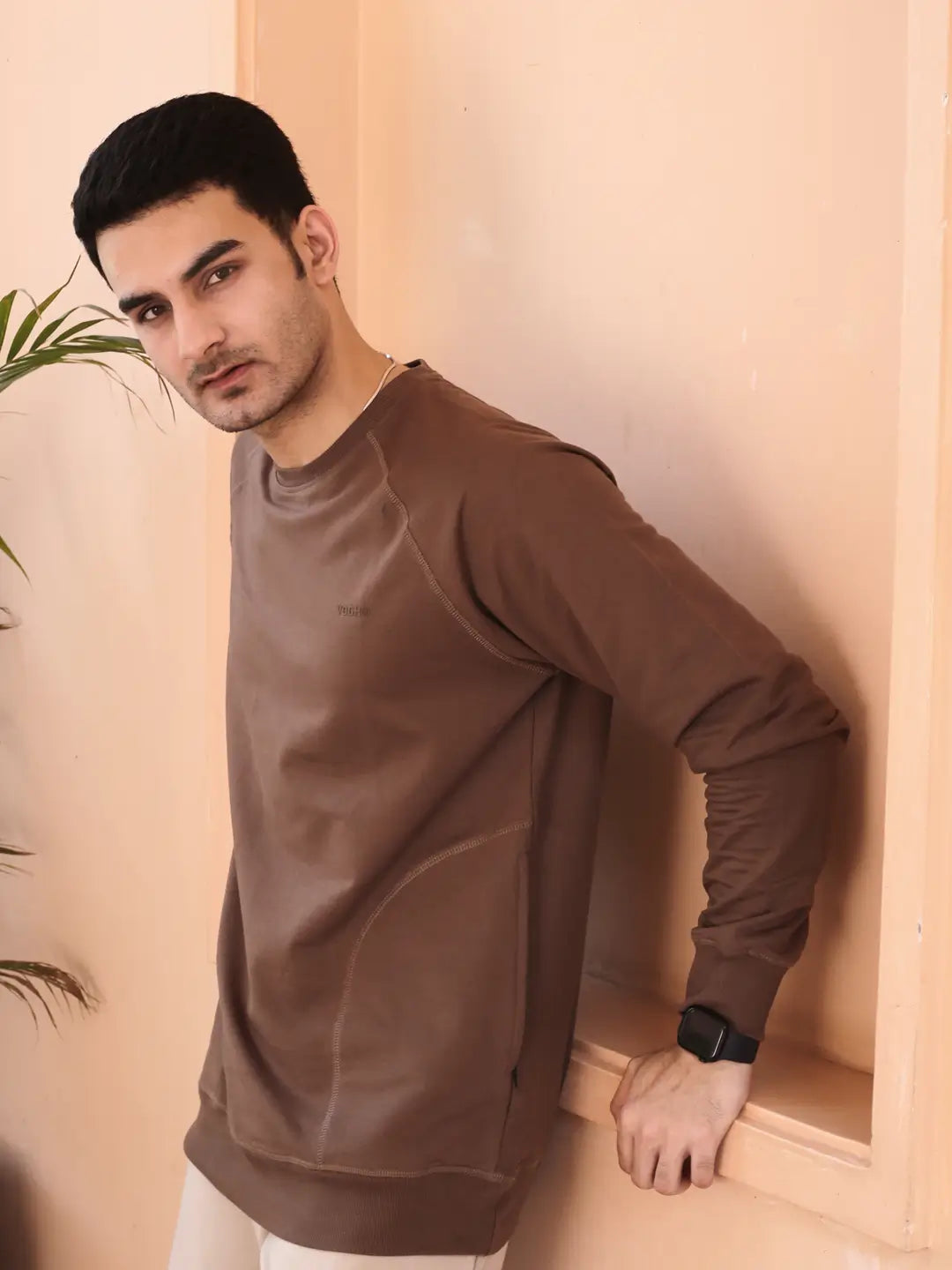 Man wearing a brown sweatshirt standing against a beige wall