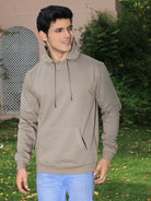 Man wearing a grey hoodie and blue jeans standing outdoors with greenery in the background