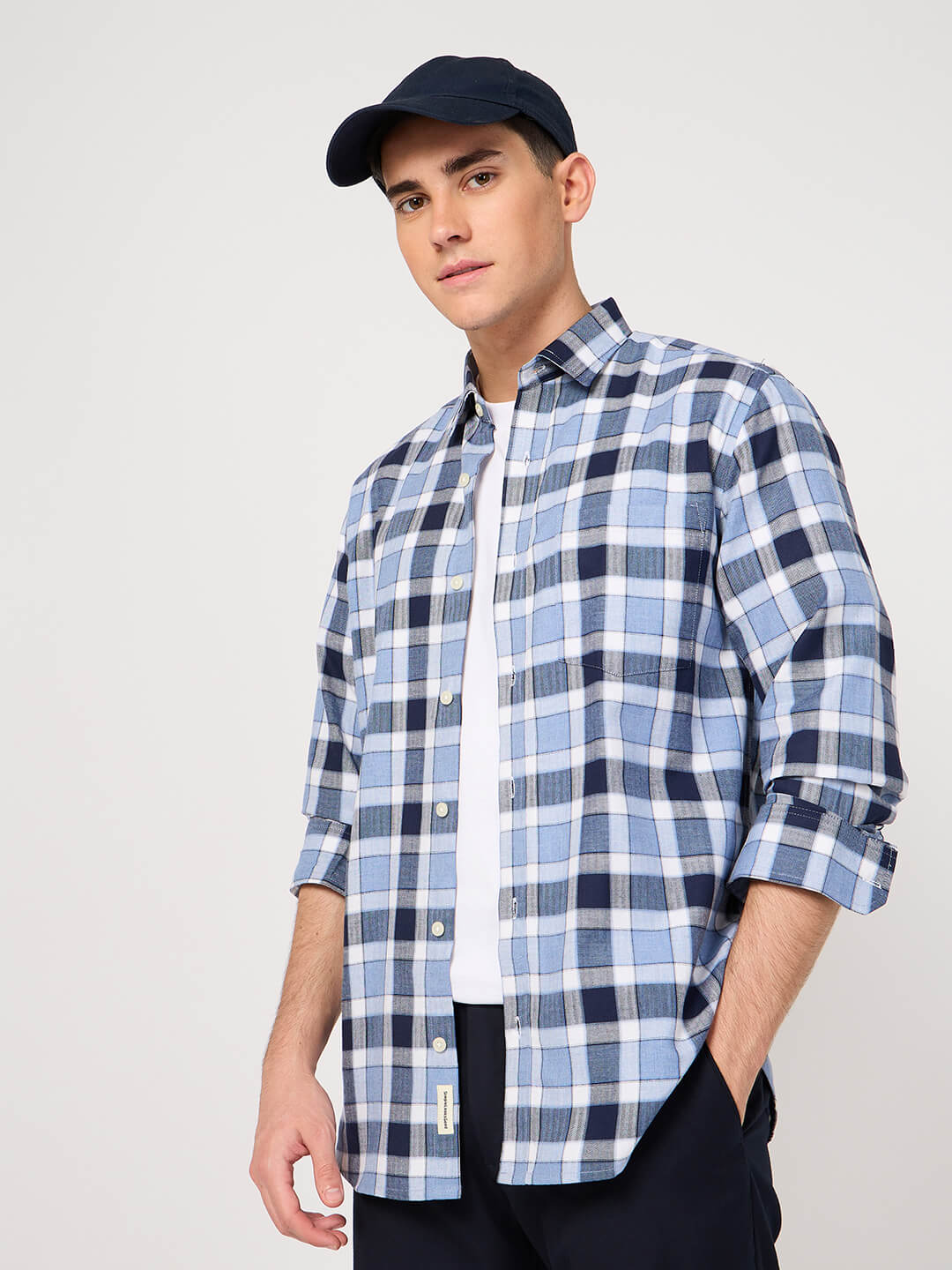Man wearing a blue and white checkered shirt with rolled-up sleeves and a navy cap on a light gray background