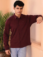 Man wearing a maroon shirt and light blue jeans standing indoors with a plant in the background
