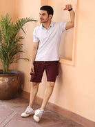 Man in a light gray polo shirt and maroon shorts standing against a beige wall