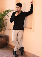 Man in black cotton shirt and beige pants standing against a wall with a plant in the background